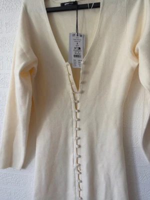 Vit lång stickad klänning Gina Tricot - Långärmad vit stickad klänning från Gina Tricot med djup v-ringning och dekorativa knappar längs framsidan. Klänningen har en figurnära passform och är tillverkad i mjukt material som passar perfekt för kyligare dagar.