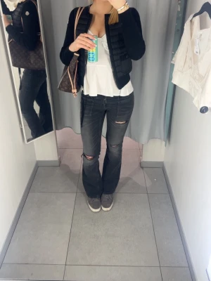 Svarta bootcut jeans med slitningar - Säljer ett par svarta bootcut jeans med slitningar på båda benen. Jeansen har en tvättad look och råa hål vid knäna för en trendig vibe. De har framfickor och är tillverkade i stretchigt denimtyg som sitter snyggt på kroppen.