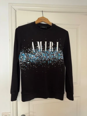 Svart sweatshirt från Amiri - Svart sweatshirt från Amiri med stort vitt logotryck och blå/vita färgstänk över bröstet. Tröjan har rund halsringning och långa ärmar. Perfekt för dig som gillar streetwear och vill sticka ut med en unik design.