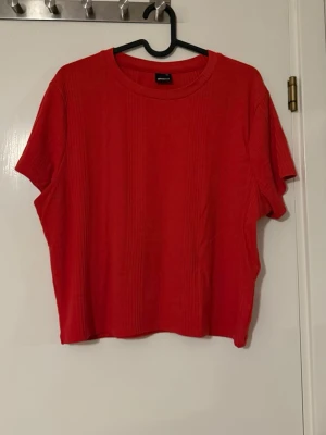 Röd ribbad topp från Gina Tricot - En snygg röd ribbad topp från Gina Tricot med rund halsringning och korta ärmar. Toppen har en loose passform och är tillverkad i ett mjukt material som känns skönt mot huden. Perfekt att styla med jeans eller kjol för en trendig look. Mindre i storlek ca M/L. 