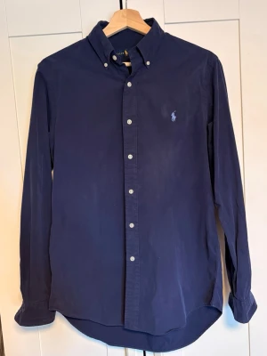 Mörkblå skjorta från Ralph Lauren - Mörkblå Polo Ralph Lauren skjorta i storlek small. Klassisk button down-modell med broderad ljusblå logga på bröstet. Funkar lika bra uppklatt som casual till jeans eller chinos. Svårt att fånga färgen på bilderna, ser ut som på första bilden men aningen mörkare. Bra skick och skönt material, säljs då den inte används lika mycket längre. Hör gärna av dig vid frågor eller om du vill ha fler bilder Nypris ligger på 1800
