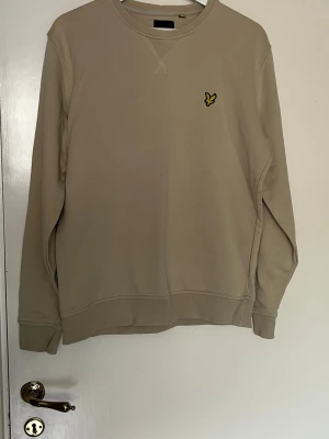 Beige sweatshirt från Lyle & Scott - Snygg beige sweatshirt från Lyle & Scott med klassisk rund halsringning och gul logga på bröstet. Tröjan har ribbade muddar vid ärmslut och nederkant. Storlek L