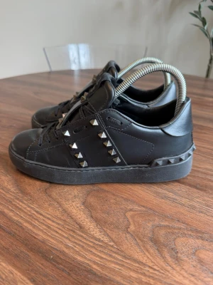 Svarta Valentino Rockstud sneakers - Snygga svarta sneakers från Valentino med ikoniska Rockstud-nitar i silver längs sidorna och bak på sulan. Skorna är i läder med svarta skosnören och har en chunky svart sula. Perfekta för dig som vill sticka ut med en edgy och lyxig stil.