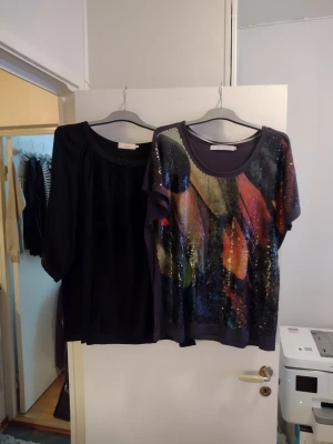 Två toppar, en svart och en glittrig - Säljer två snygga toppar. Ena är helsvart med rund hals och trekvartsärm, den andra har ett färgglatt abstrakt mönster i lila, orange, rött och blått med glittriga paljetter och kort ärm. Båda är i mjukt material och har avslappnad passform. Zizzi & Zj denim by Zizzi