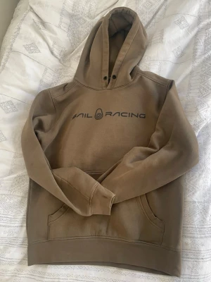 Beige hoodie från Sail Racing - Snygg beige hoodie från Sail Racing med svart logga tryckt på bröstet. Klassisk modell med huva, känguruficka och ribbade muddar. Perfekt för en avslappnad och sportig stil.