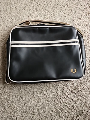 Svart axelväska från Fred Perry - Snygg svart axelväska från Fred Perry med vita detaljer och dubbla ränder framtill. Väskan har en klassisk rektangulär form, justerbar axelrem och är tillverkad i ett glansigt konstlädermaterial. Guldigt Fred Perry-logo på framsidan ger en sportig touch. Helt ny, knappt använd.