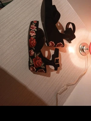 Svarta sandaler med rosbrodyr - Säljer ett par svarta sandaler med platåsula och broderade röda rosor på sidorna. Sandalerna har en justerbar rem med guldfärgat spänne och öppen tå. Materialet ser ut att vara mockaimitation och sulan är i gummi. Perfekta för dig som vill sticka ut lite extra.