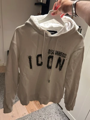 Vit hoodie Dsquared2 ICON - Säljer en vit hoodie från Dsquared2 med stor svart 'ICON'-text och Dsquared2-logga framtill. Hoodien har huva med dragsko och ribbade muddar. Materialet känns mjukt och skönt, perfekt för en avslappnad streetwear-look. Storlek M men passar mer som S/M.