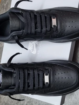 Nike Air Force 1 Low helsvart - Nike Air Force 1 Low i helsvart läder med klassisk siluett och perforerad tåbox. Skorna har svarta skosnören, metallbricka med AF1-logga och diskret Nike-märke på plösen. Snygg och tidlös sneaker som passar till det mesta.