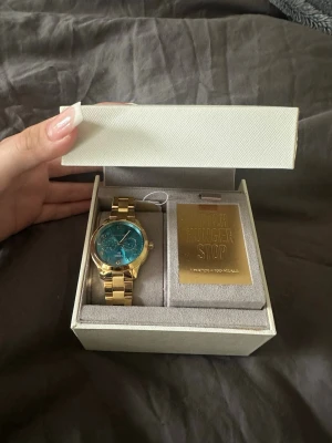 Guldfärgad klocka Michael Kors - Snygg guldfärgad klocka från Michael Kors med blå urtavla och metallarmband. Kommer i originalask med etikett och märkning 'Watch Hunger Stop'. Perfekt accessoar för dig som vill sticka ut med stilren design och exklusiv känsla.