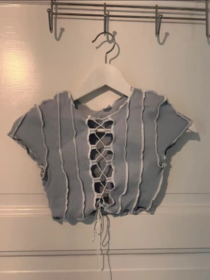 Ljusblå croppad topp med snörning - Trendig ljusblå croppad topp med korta ärmar och ribbad struktur. Toppen har råa kanter och en snygg snörning framtill som ger en edgy vibe. Perfekt för dig som vill sticka ut med en unik detalj. Materialet känns mjukt och stretchigt. Passar bra med bland annat ett par jeans.