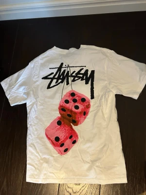 Vit Stussy t-shir S - Vit t-shirt från Stüssy med svart logga på bröstet och stort tryck av två rosa tärningar på ryggen. Klassisk passform och rund hals. Perfekt för dig som gillar streetwear och vill sticka ut med en unik design. Använd 1 gång endast.