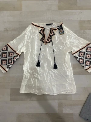 Vit blus med broderade ärmar och snörning - Vit blus med broderade detaljer i svart, rosa och orange på ärmarna och vid halsen. Blusen har snörning med tofsar framtill och trekvartsärmar. Tillverkad i ett glansigt, lätt material som ger en lyxig känsla.