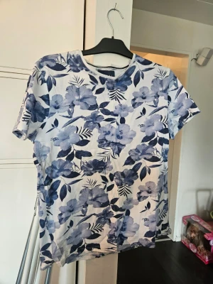 Blommig t-shirt från Bondelid - Snygg t-shirt från Bondelid med blått och vitt blommönster. T-shirten har rund halsringning och korta ärmar. Tillverkad i mjuk bomull som känns skön mot huden. Perfekt för dig som gillar en fräsch och somrig stil.