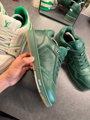 Gröna Dior B27 sneakers i läder - Snygga gröna Dior sneakers i lågt modell med ovandel i läder och diskret Dior-logga på sidan. Skorna har vita detaljer på sulan och klassisk snörning. Perfekta för dig som vill sticka ut med exklusiva sneakers och gillar streetwear-vibe.