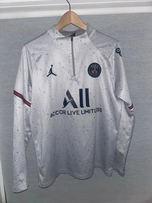 Vit PSG träningströja Jordan - Snygg vit långärmad träningströja från PSG och Jordan med svart stänkmönster, halv dragkedja och klubbmärke på bröstet. Tröjan har blå och röda detaljer på ärmarna och tryck på både fram- och baksida. Perfekt för fotbollsträning eller sport.