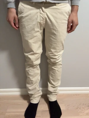 Beige chinos med smal passform från NN07 - Snygga beige chinos med smal passform och klassiska fickor fram och bak. Byxorna har bälteshällor och stängs med knapp och dragkedja. Perfekta för en clean och stilren look. Tillverkade i mjukt bomullstyg som känns skönt mot huden.