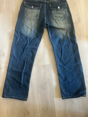 True Religion blå jeans med kontrastsöm - Säljer ett par blå jeans från True Religion med raka ben och tydliga kontrastsömmar. Bakfickorna har lock och dekorativa vita sömmar samt knappstängning. Klassisk denimlook med snygg tvätt och detaljerad etikett bak.
