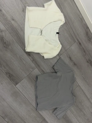 Vit fluffig bolerotopp & grå croppad topp - Två trendiga toppar: en vit fluffig bolerotopp med v-ringning och dekorativ knäppning framtill, samt en enkel grå croppad topp med korta ärmar. Perfekta att styla lager på lager eller var för sig för en cool look.