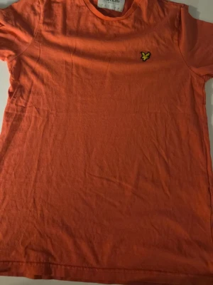 Orange t-shirt från Lyle & Scott - Råsa orange t-shirt från Lyle & Scott med klassisk passform och korta ärmar. T-shirten har en broderad gul logotyp på bröstet och är tillverkad i mjuk bomull. Perfekt för en avslappnad och stilren look.