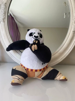 Kung Fu Panda mjukisdjur - Säljer ett mjukisdjur som föreställer Po från Kung Fu Panda. Gosedjuret är svartvitt med gröna broderade ögon och har på sig beige byxor med röda och gula detaljer. Tillverkad i mjukt plyschmaterial och har broderade tassar.