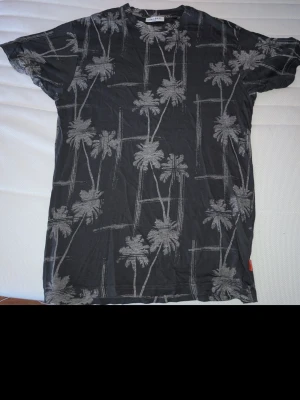 Svart t-shirt med palmtryck - Snygg svart t-shirt med grått palmtryck över hela plagget. Klassisk passform och korta ärmar, perfekt för en avslappnad stil. Tillverkad i mjukt material som känns skönt mot huden. Perfekt för dig som gillar streetwear och somriga vibes.
