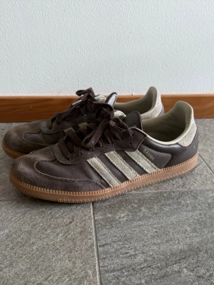 Adidas Samba bruna sneakers - Klassiska Adidas Samba sneakers i brunt läder och mocka med beige detaljer och de ikoniska tre ränderna på sidan. Yttersulan är i gummi och skorna har låga skaft. Perfekta för dig som gillar retrostil och streetwear. Storlek 44