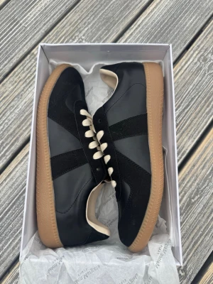 Maison Margiela svarta sneakers - Maison Margiela sneakers i svart läder och mocka med beige snörning och klassisk gummisula. Snygg retrostil med låg profil och diskreta detaljer. Perfekta för dig som gillar minimalistisk och stilren design.