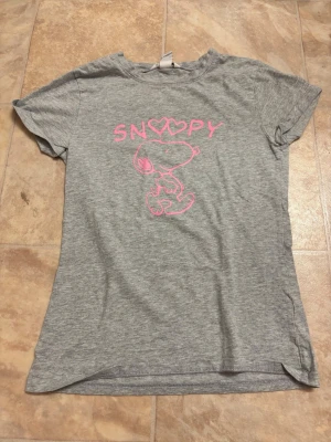 Grå Snoopy t-shirt med rosa tryck - Snygg grå t-shirt med ett rosa Snoopy-tryck och text framtill. T-shirten har korta ärmar och en klassisk passform. Perfekt för dig som gillar gulliga och avslappnade plagg med en lekfull touch.