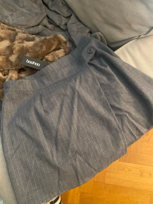 Grå randig omlottkjol  - Snygg grå kjol från Boohoo med diskreta vita ränder. Stängs med en knapp på insidan av kjolen och en synlig knapp på utsidan. Aldrig använd. Storlek 38 på lappen, men jag upplever storleken som 34-36.