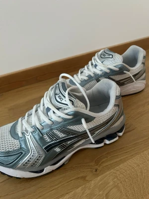 Asics vita och silver sneakers - Snygga sneakers från Asics i vitt och silver med mesh-ovandel och detaljer i syntet. Skorna har klassisk snörning och låg profil, perfekt för en sportig och trendig look. Sulan är vit med svarta och silvriga inslag. Perfekta för dig som gillar retrostil.