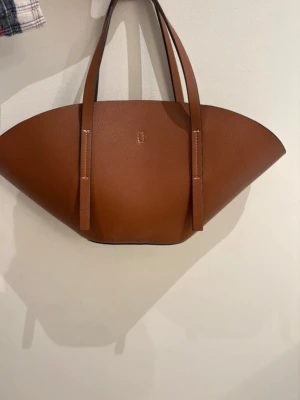 Brun shoppingväska från Gina Tricot - Snygg shoppingväska i brunt konstläder från Gina Tricot. Väskan har en unik bågformad design, dubbla axelband och ett stilrent metallspänne på insidan. Perfekt för dig som vill ha en trendig och rymlig väska till vardagen.