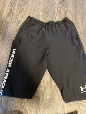 Svarta träningsbyxor Under Armour - Snygga svarta träningsbyxor från Under Armour med vitt logotryck längs benet och liten logga vid benöppningen. Byxorna har resår i midjan och är gjorda i ett lätt, snabbtorkande material som passar perfekt till gymmet eller löpning.