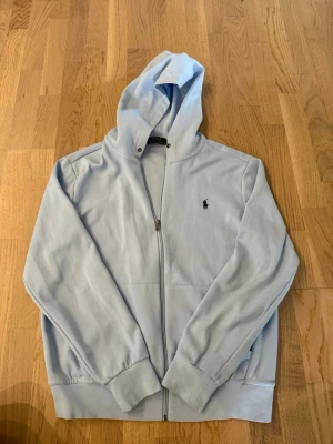Ljusblå hoodie från Polo Ralph Lauren - Snygg ljusblå hoodie med dragkedja från Polo Ralph Lauren. Klassisk design med huva och det ikoniska broderade logotypen på bröstet. Tillverkad i mjukt bomullsmaterial och har ribbade muddar vid ärmslut och nederkant.