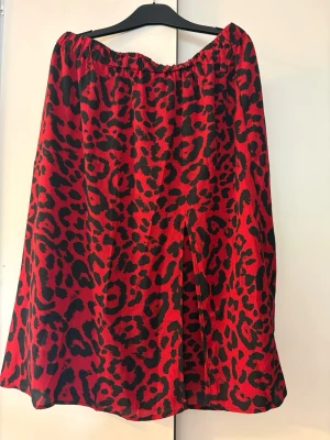 Röd leopardmönstrad halv lång kjol - Cool röd långkjol med svart leopardmönster och lätt utställd passform. Kjolen har resår i midjan för extra komfort och en liten slits framtill. Perfekt statement-plagg för dig som vill sticka ut.