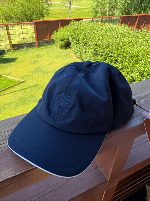 LP cap - Snygg mörkblå keps från Lyle & Scott med broderad logga framtill. Kepsen har böjd skärm och justerbar passform baktill. Insidan har ett rutigt tygfoder och brunt svettband. Perfekt accessoar för en sportig och stilren look.