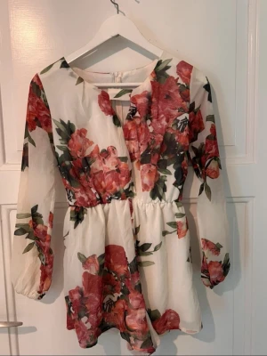 Vit blommig playsuit med långa ärmar - Söt vit playsuit med stora blommiga mönster i rött och grönt. Playsuit har långa ärmar, markerad midja och rund halsringning. Perfekt för vår och sommar med sitt luftiga material och romantiska look. Notera tredje bilden, lätt att sy fast.