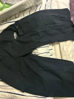 Svarta cargobyxor från Ellesse - Svarta cargobyxor från Ellesse med klassisk logga på benet. Byxorna har flera fickor, bland annat en med lock och knapp, samt dragsko i midjan för skön passform. Perfekta för en avslappnad streetwear-look.