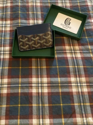  Blå korthållare. - Snyggt blått kortfodral med det klassiska mönstret i vitt och beige. Tillverkat i canvas och läder, perfekt för kort och sedlar. Stilren och lyxig accessoar för dig som gillar exklusiva detaljer.