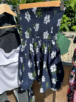Blommig marinblå klänning Hollister - Man kan knyta en rosett med bandet. Söt marinblå kortklänning från Hollister med vita blommor och gröna detaljer. Klänningen är axelbandslös med smock upptill och har en luftig kjol. Perfekt för varma dagar och sommarhäng.