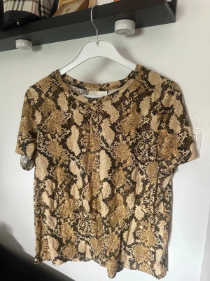  t-shirt med ormmönster - Cool t-shirt med beige och brunt ormmönster. T-shirten har rund halsringning och korta ärmar, perfekt för dig som vill sticka ut med en trendig look. Materialet känns mjukt och lätt, passar bra till jeans eller kjol.