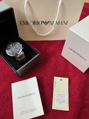 Emporio Armani blå-silver klocka - Stilren klocka från Emporio Armani med blå urtavla och silverfärgad länk i metall. Klockan har kronograf-funktion, datumvisning och klassiska guldfärgade index. Levereras i originalbox med certifikat och manual.
