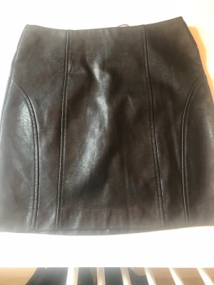 Svart kort kjol i skinnimitation, helt ny - Snygg svart kort kjol från Stradivarius i skinnimitation. Kjolen har en stilren design med markerade sömmar framtill och en tight passform. Perfekt att matcha med en stickad tröja eller en cool t-shirt. Helt ny.