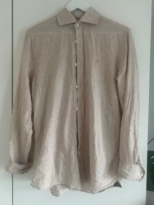 Beige linneskjorta från Morris - Snygg beige skjorta i linne med klassisk krage och knappar framtill. Skjortan har ett diskret broderat emblem på bröstet och långa ärmar med manschett. Perfekt för en clean och avslappnad stil.
