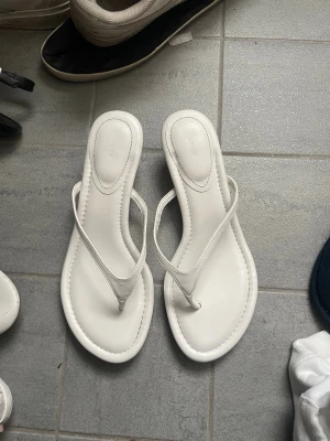 Vita sandaletter med låg klack från Nelly - Säljer ett par stilrena vita sandaletter från Nelly med tunna remmar och låg klack. Sandalerna har en klassisk flip-flop design och är tillverkade i ett slätt syntetmaterial. Perfekta för dig som vill ha en enkel men trendig look. Dom är testade ute en gång och är i storlek 38.
