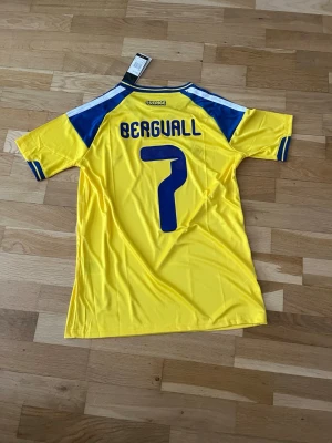 Sverige matchtröja Adidas gul/blå - Säljer en officiell Sverige matchtröja från Adidas i gult med blå detaljer och nummer 7 samt namnet 'Bergvall' på ryggen. Tröjan har korta ärmar, rund hals och broderad landslagssköld på bröstet. Tillverkad i lätt och ventilerande material.