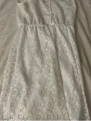 Vit spetsklänning med rund hals - Söt vit klänning i spets med rund halsringning och ärmlös design. Klänningen har ett fint spetsmönster över hela plagget och markerad midja för en feminin siluett. Perfekt för sommarens alla tillfällen.