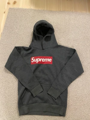 Mörkgrå Supreme hoodie - Mörkgrå hoodie från Supreme med den klassiska röda boxloggan på bröstet. Tröjan har huva och en stor magficka framtill. Tillverkad i mjukt bomullsmaterial och har ribbade muddar vid ärmslut och nederkant. Perfekt för dig som gillar streetwear och vill sticka ut.