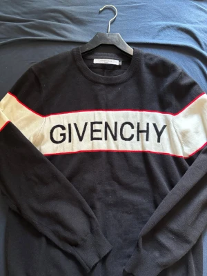 Svart stickad tröja från Givenchy - Snygg svart stickad tröja från Givenchy med vit panel och röd kant över bröstet och armarna. Stor Givenchy-logga framtill. Rund halsringning och långa ärmar. Perfekt för dig som gillar streetstyle och exklusiva märken.