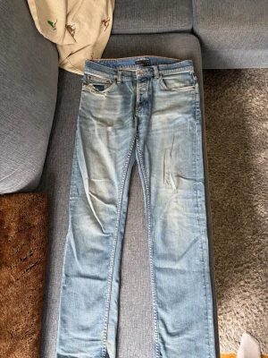 Ljusblå Nudie Jeans 31/32 - Säljer ett par ljusblå jeans från Nudie Jeans i storlek 31/32. Jeansen har klassisk femficksmodell, raka ben och slitna detaljer för en avslappnad look. Tillverkade i denim med kontrastsömmar och metallknappar.nypris: 1800kr